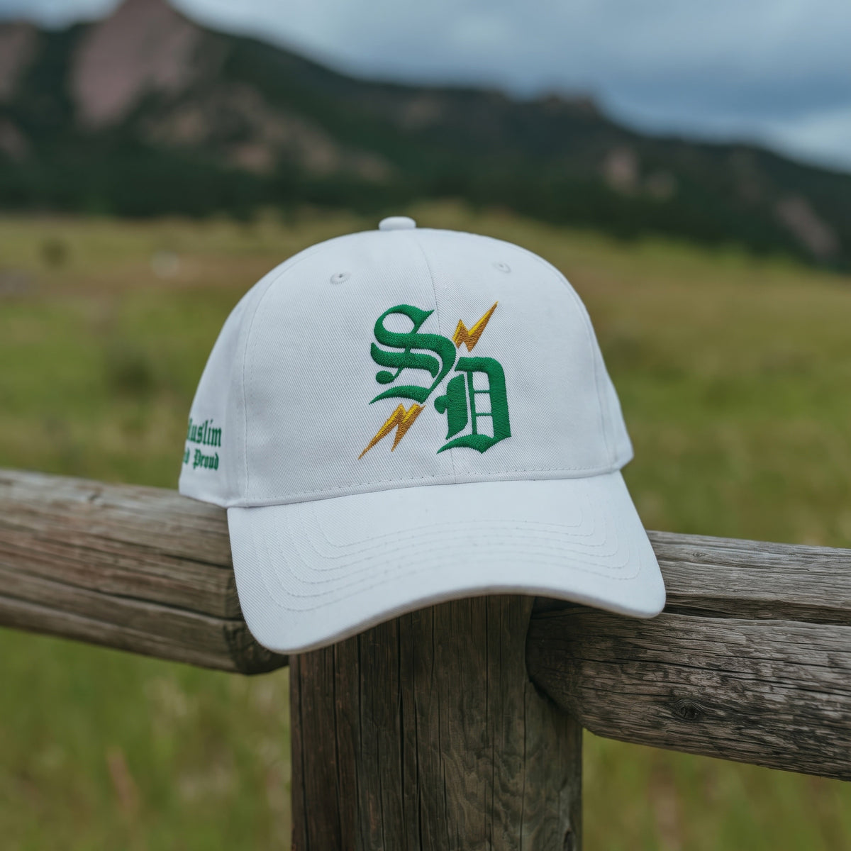 Street Dawah Apparel white 5-panel structured cap with green and gold embroidered logo on a wooden fence outdoors.